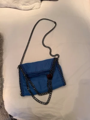 Blå Stella McCartney liknande axelväska med kedja - Snygg blå axelväska med silverfärgad kedjerem och dekorativa kedjedetaljer längs kanterna. Väskan har en kuvertformad design och stängs med magnetknapp. Perfekt för dig som vill ha en cool accessoar till outfiten.