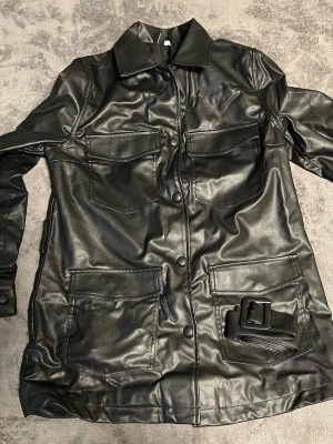 Svart lång skinnjacka med bälte - Snygg svart skinnjacka i längre modell med fyra stora fickor framtill och tillhörande bälte med stor spänne. Jackan har klassisk krage, knappar längs hela framsidan och raka ärmar. Perfekt för dig som vill ha en edgy och stilren look. Inte i äkta skinn, polyester.