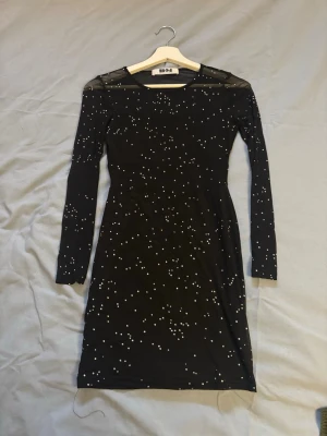 Svart glittrig meshklänning DM - Säljer en svart kort klänning från DM med glittriga detaljer och meshpartier. Klänningen har långärmade ärmar och en kroppsnära passform. Perfekt för dig som vill sticka ut med en trendig look.
