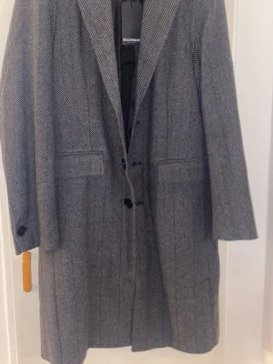 Grå mönstrad kappa från Bolongaro - Stilren grå kappa från Bolongaro med klassiskt fiskbensmönster. Kappan har två framfickor, knappar framtill och en rak passform. Perfekt för dig som gillar tidlös design och vill ha en snygg ytterrock till höst och vinter.