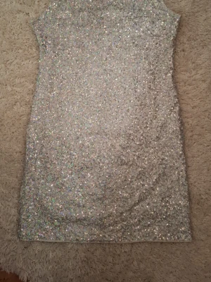 Glittrig silver kortklänning - Supersnygg glittrig kortklänning i silver med paljetter över hela klänningen. Klänningen är ärmlös och har en öppen rygg med rundad öppning. Perfekt för dig som vill sticka ut och glänsa på nästa fest eller bal.