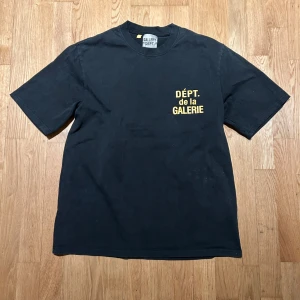 Svart t-shirt från Gallery Dept. - Cool svart t-shirt från Gallery Dept. med gul text 'DÉPT. de la GALERIE' tryckt både fram och bak. Klassisk passform och rund hals. Tillverkad i mjuk bomull, perfekt för dig som gillar streetwear och vill sticka ut med en grafisk look. Aldrig använd. ⚠️1:1 ⚠️