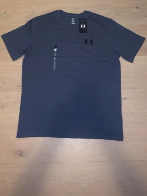 Mörkgrå Under Armour t-shirt - Snygg mörkgrå t-shirt från Under Armour med klassisk logga tryckt på bröstet. T-shirten har rund halsringning och korta ärmar. Tillverkad i ett mjukt och funktionellt syntetmaterial som passar perfekt till träning.