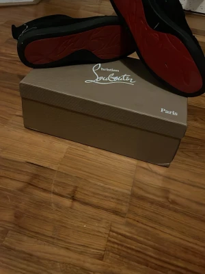 Svarta Christian Louboutin sneakers - Snygga svarta sneakers från Christian Louboutin med ikonisk röd sula. Skorna har klassisk rund tå, snörning och är tillverkade i ett material som ser ut som mocka. Perfekta för dig som vill sticka ut med stilrena detaljer. Höger sko är helt trasig men går att använda fint ändå, jag tycker inte att det märks när man använder dom. box och dustbag fås med vid rätt pris och enkel affär. 