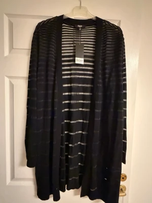 Svartvit randig kofta Slim Factor - Snygg lång svart kofta från Slim Factor med tunna vita horisontella ränder. Öppen framtill och tillverkad i ett lätt stickat material som ger en avslappnad look. Perfekt att slänga över en t-shirt eller topp för extra stil.