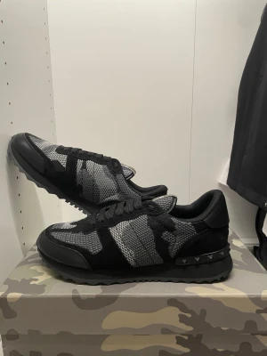 Valentino Rockrunner sneakers svart/grå - Säljer ett par Valentino Rockrunner sneakers i svart och grått med snyggt camouflage-mönster. Skorna har snörning, rund tå och platt sula. Ovandelen är i mesh och mocka med coola nitar på hälen. Perfekt för dig som gillar streetstyle och exklusiva detaljer.