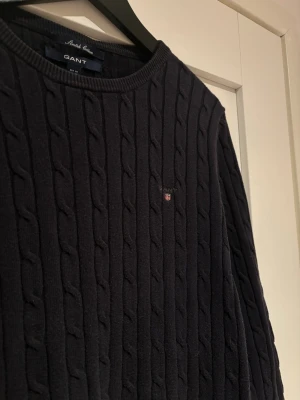 Mörkblå kabelstickad tröja GANT - Kabelstickad tröja från GANT i mörkblått med rund halsringning och diskret logga broderad på bröstet. Tröjan är i nyskick, inga defekter, hål eller fläckar!