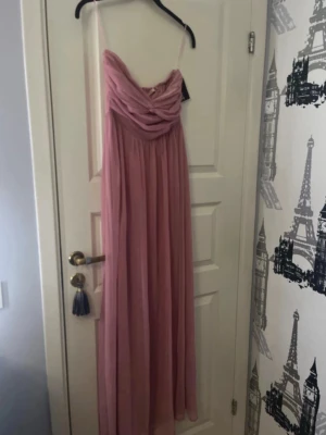 Lång rosa klänning från NLY Eve - Romantisk långklänning i mjuk rosa nyans från NLY Eve. Klänningen har draperad överdel, tunna axelband som går att ta bort om man vill, och ett flytande fall i tyget. Perfekt för speciella tillfällen och har en feminin siluett med vackra detaljer.