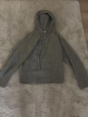 Grå stickad hoodie med huva - Mysig grå stickad hoodie med huva och dragsnören. Tröjan har breda ribbstickade muddar vid ärmslut och nederkant, samt en avslappnad passform. Perfekt för en chill och stilren look.