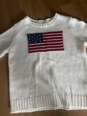 Vit stickad tröja med USA-flagga - Vit stickad tröja med lång ärm och ribbade muddar. På bröstet finns en stor amerikansk flagga i blått, rött och vitt. Tröjan har rund halsringning och är gjord i mjukt material, perfekt för en avslappnad och cool stil.