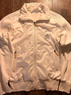 Vit Adidas track jacket - Snygg vit track jacket från Adidas med klassiska tre ränder längs ärmarna och broderad logga på bröstet. Dragkedja framtill och två fickor med dragkedja. Perfekt för en sportig och avslappnad stil.