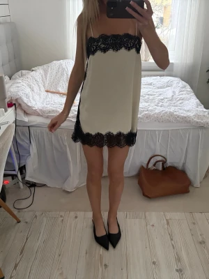 Krämfärgat klänning med spetsdetaljer - Snyggt nattlinne i krämvitt med tunna axelband och svarta spetsdetaljer upptill och nedtill. Kort modell med rak passform och elegant kontrast mellan tyg och spets. Perfekt för en chic och bekväm kväll hemma.