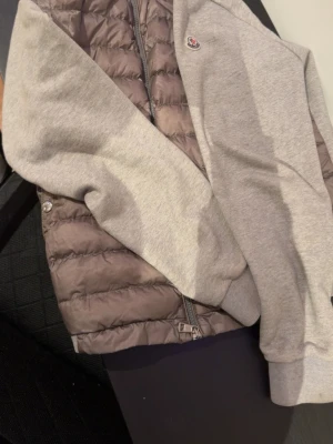 Grå quiltad cardigan jacka Moncler - Snygg cardigan jacka från Moncler med quiltad, mörkgrå kropp och ljusgrå ärmar i sweatshirtmaterial. Jackan har dragkedja framtill och en liten Moncler-logga på bröstet. Perfekt för dig som vill ha en sportig men stilren look.