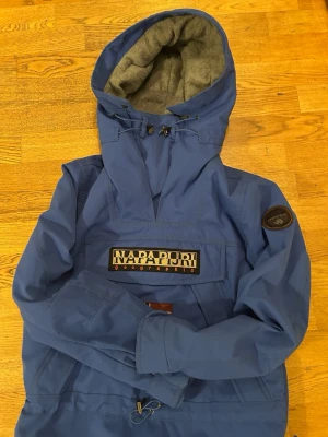 Blå skidjacka från Napapijri - Snygg blå skidjacka från Napapijri med stor huva och fleecefoder. Jackan har en stor logotyp framtill, patch på ärmen och praktisk dragkedja vid halsen. Perfekt för blåsiga dagar och streetstyle.