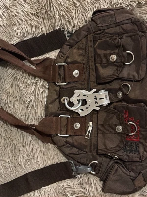 Brun GG&L ryggsäck med detaljer - Cool brun ryggsäck från George Gina & Lucy med flera fickor, silvriga metalldetaljer och ett stort GG&L-hänge framtill. Väskan har justerbara axelband, dragkedjor och tryckknappar. Materialet är slitstark polyester med camouflageinspirerat mönster.