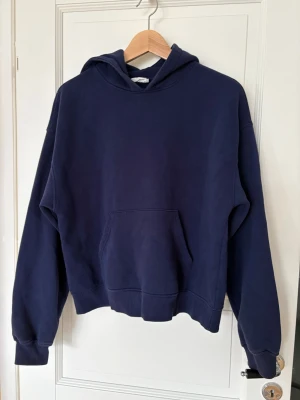 Mörkblå hoodie med magficka - En enkel och stilren mörkblå hoodie med stor huva och rymlig magficka. Hoodien har ribbade muddar vid ärmslut och nederkant, samt en avslappnad passform. Perfekt för dig som gillar en clean och bekväm look. Materialet känns mjukt och skönt mot huden.