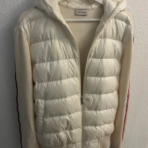 Moncler Cardigan - Snygg vit pufferjacka från Moncler med stickade ärmar och quiltad front. Jackan har huva, dragkedja och Moncler-logga på ärmen. Röd, vit och blå rand längs ärmarna. Perfekt för dig som vill ha en clean och exklusiv look. Kvitto finns! 