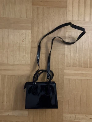 Svart lackad handväska med axelrem - Snygg svart handväska i lackat material med både handtag och lång axelrem. Väskan har en fyrkantig form och stängs med dragkedja. Perfekt för dig som vill ha en stilren och trendig accessoar till din outfit. Lite sönder vid sidan