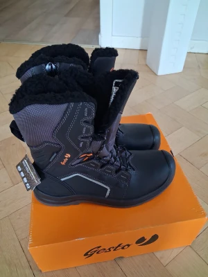 Svarta vinterstövlar Gesto Tor strl 40 - Svarta vinterstövlar från Gesto, modell Tor, med snörning och fluffigt svart foder. Stövlarna har robust platt sula, förstärkta detaljer och reflekterande inslag. Utsidan är i slitstark syntet och skinn, perfekt för kalla dagar. Snyggt orange logotyptryck på sidan.... ny pris ca 2000 kr
