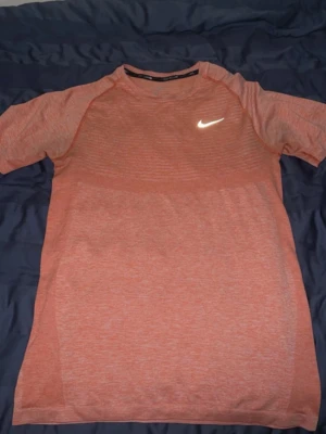 Nike t-shirt dri-fit - Snygg rosa t-shirt från Nike med diskret randigt mönster och klassisk vit logga på bröstet. T-shirten har korta ärmar och är tillverkad i ett lätt och ventilerande material, perfekt för träning eller chill vardagsstil.