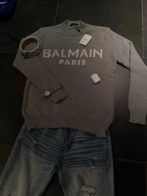 Grå stickad tröja från Balmain - Snygg grå stickad tröja från Balmain Paris med vit logotyp framtill. Tröjan har rund halsringning och ribbstickade muddar vid ärmslut och nederkant. Perfekt för dig som gillar exklusiva och trendiga plagg med tydlig logga.