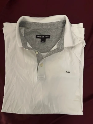 Vit piké från Michael Kors - Snygg vit pikétröja från Michael Kors med grå detaljer på kragen och insidan av knäppningen. Klassisk passform med korta ärmar och diskret MK-logga broderad på bröstet. Perfekt till jeans eller shorts för en clean och stilren look.