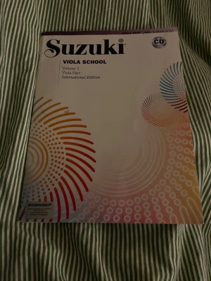 Suzuki Viola School Vol 1 + CD - Suzuki Viola School Volume 1, International Edition. Notbok för viola med tillhörande CD. Omslaget är vitt med färgglada cirkelmönster i orange, rött, blått och gult. Perfekt för dig som vill lära dig spela viola med Suzuki-metoden.