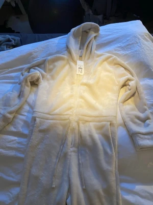 Krämvit fleece onepiece med huva - Supermjuk och mysig krämvit onepiece i fleece med dragkedja framtill, huva och dragsko i midjan. Perfekt för chill dagar hemma. Två fickor framtill och långa ärmar för extra komfort. Helt ny med lapp kvar.