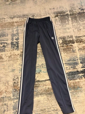 Mörkblå Adidas mjukisbyxor - Säljer ett par mörkblå Adidas mjukisbyxor med vita sidoränder och dragkedjefickor. Byxorna har elastisk midja och den klassiska Adidas-loggan på vänster sida. Perfekta för chill eller träning.