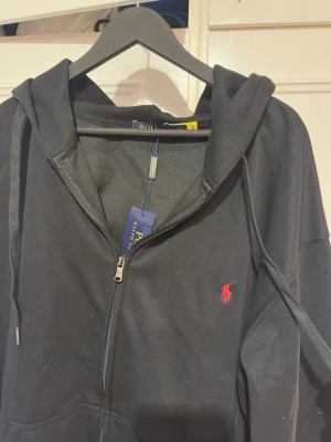 Svart hoodie från Polo Ralph Lauren - Svart hoodie från Polo Ralph Lauren med dragkedja och huva. Klassisk röd broderad logga på bröstet och snörning i huvan. Tillverkad i mjuk bomull, perfekt för en chill och stilren look.