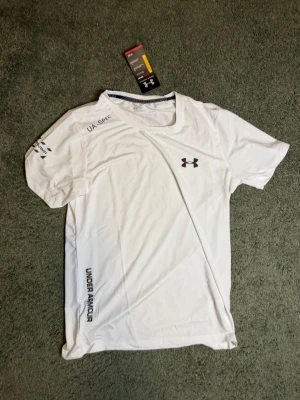 Vit sport-t-shirt från Under Armour - Snygg vit sport-t-shirt från Under Armour med korta ärmar och rund hals. T-shirten har tryckt logga på bröstet och textdetaljer på ärm och sida. Tillverkad i lätt och ventilerande funktionsmaterial, perfekt för träning.