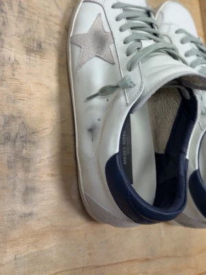 Golden Goose vita sneakers med stjärna - Golden Goose sneakers i vitt läder med grå mockastjärna på sidan, grå skosnören och mörkblå detalj vid hälen. Insidan är fodrad med beige tyg och sulan är vit. Sidan har texten GGBD/STAR. Klassisk low-top modell med retrovibe. Skick 8,5/10 knappt använda. Skriv vid frågor!🙏🏻