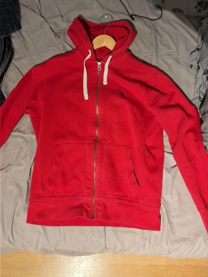 Ralph Lauren Hoodie - Röd hoodie med dragkedja från Polo Ralph Lauren. Klassisk design, två framfickor och broderad marinblå logga på bröstet. Mjuk bomullskänsla och ribbade muddar vid ärmslut och nederkant. 