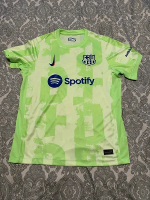 Barcelona Nike matchtröja neongrön - Snygg Barcelona matchtröja från Nike i neongrönt med klubbmärke och Spotify-logga på bröstet. Tröjan har korta ärmar, rund hals och tryckta detaljer i blått och rosa. Perfekt för fotbollsträning eller att bära till vardags.