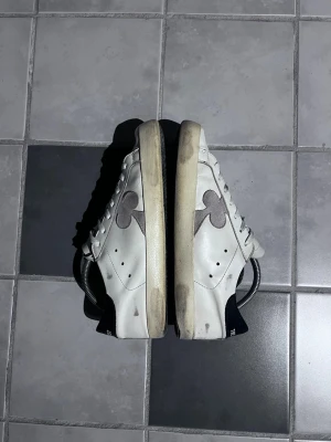 Vita sneakers från The Gallerist - Snygga vita sneakers från The Gallerist med grå klöverdetalj på sidan och svart hälparti. Skorna har klassisk låg profil, vita skosnören och ovandel i läder. Perfekta för en stilren och trendig look. Perfekt till sommaren och våren. Skorna har redan en lite distressed look, som ska se ut så. Den enda skadan är på hälen, där sulan har slipats ner, men det läcker inte inte vatten eller liknande. Hör av er vid eventuella frågor, priset kan diskuteras. Nypris då var 2400. 