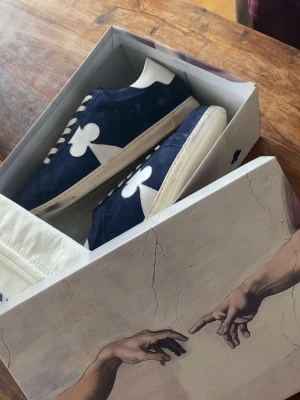 The Gallerist sneakers  - Snygga The Gallerist  sneakers i mörkblå mocka med vita detaljer och den ikoniska blomman på sidan. Vita skosnören och sula ger en clean kontrast. Perfekta för dig som vill sticka ut med stilrena och unika sneakers. (Använda 2 gånger) säljer bga. För stor storlek då jag har 42 och dem är 43