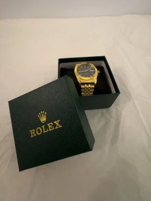  klocka med svart urtavla - Snygg guldfärgad klocka  med svart urtavla och tydliga index. Klockan har en klassisk länk i metall och levereras i original grön -box. Perfekt accessoar för dig som vill sticka ut med stil och elegans.