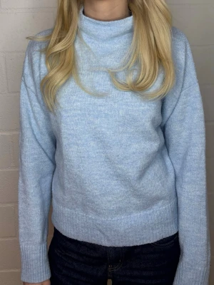 Ljusblå stickad polotröja - Mysig ljusblå stickad tröja med hög krage och långa ärmar. Tröjan har en rak passform och ribbade muddar vid ärmslut och nederkant. Perfekt för kyliga dagar och enkel att matcha med jeans eller kjol.