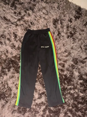 Palm Angels svarta mjukisbyxor - Säljer ett par svarta mjukisbyxor från Palm Angels med färgglada ränder i grönt, gult och rött längs sidorna. Byxorna har resår i midjan och logga broderad på vänster sida. Perfekta för en chill och trendig streetwear-look.