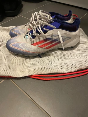 Adidas f50 elite ag fotbollsskor - Snygga Adidas f50 ag elite fotbollsskor i vitt med röda och blå detaljer. Skorna har låg vikt, snörning och AG-dobbar för spel på konstgräs. Perfekta för dig som vill ha fart på planen. Använda max 3 gånger på konstgräs. Nypris ca 2-3 tusen