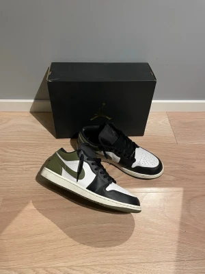 Nike air Jordan 1 Low svart/vit/oliv - Säljer nu ett par snygga Air Jordan 1 Low sneakers i svart, vitt och olivgrönt läder. Klassisk siluett med svart snörning, vit tåbox och olivgröna detaljer på häl och Swoosh. Perfekta för dig som gillar streetwear. Det är storlek 47 men de är lite små i storlek så skulle säga att de passar 46 också. Hör av dig vid eventuella frågor☺️