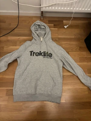 Grå Treklife hoodie med tryck - Säljer en grå hoodie från Treklife med svart tryck på bröstet där det står 'Treklife NEW YORK TRAIL'. Hoodien har en klassisk huva med dragsko och en stor magficka. Perfekt för chill dagar och enkel att matcha med jeans eller joggers.