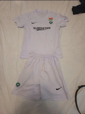 Vit fotbollsdräkt Kurdistan Nike - Säljer ett vitt fotbollsset från Nike med Kurdistan-tryck på bröstet, Kurdistans flagga och klubbmärke. Setet består av en kortärmad tröja och matchande shorts i lätt och ventilerande material, perfekt för träning eller match.