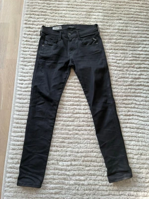 Svarta slim fit jeans från Replay - Snygga svarta slim fit jeans från Replay, modell Anbass. Klassisk femficksdesign med dragkedjegylf och diskret Replay-logga vid fickan. Jeansen har en modern passform och är tillverkade i ett stretchigt material för extra komfort. Storlek 30