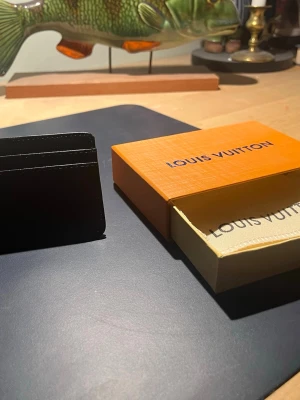 Svart Louis Vuitton kortfodral - Snyggt svart kortfodral från Louis Vuitton i slimmad design. Tillverkat i exklusivt läder med plats för flera kort. Kommer med original orange box och dustbag med Louis Vuitton-logga. Perfekt för dig som vill ha en stilren och lyxig accessoar.