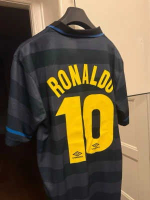 Inter Ronaldo 10 matchtröja Umbro - Säljer en klassisk Inter Milan matchtröja från Umbro med Ronaldo och nummer 10 på ryggen. Tröjan är svart med mörkblå horisontella ränder, gula tryck och blå detaljer vid kragen och ärmsluten. Perfekt för dig som älskar retro fotbollströjor! Helt ny