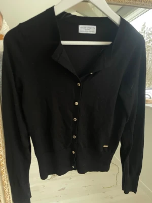 Svart kofta från Holly & White - Svart kofta med långa ärmar från Holly & White. Klassisk design med rund halsringning och knappar framtill i silverfärg. Mjuk och tunn kvalitet, perfekt att ha över en topp eller klänning. Liten metallbricka vid nederkanten för extra detalj.