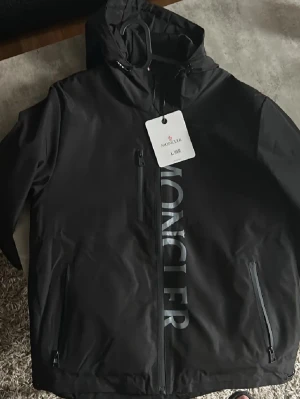 Svart vindjacka från Moncler - Snygg svart vindjacka från Moncler med stor logga längs framsidan. Jackan har huva, tre dragkedjeförsedda fickor och justerbar nederkant. Tillverkad i ett lätt, vattenavvisande material som passar perfekt för blåsiga dagar. Stilsäker och funktionell design.