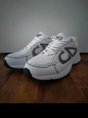 Vita Dior B30 sneakers - Snygga vita Dior B30 sneakers med grå och svarta detaljer. Skorna har en sportig siluett, mesh- och läderpaneler samt den ikoniska Dior-loggan på sidan. Perfekta för dig som gillar exklusiva och trendiga sneakers.skriv till mig för bilder eller om du är intresserad!