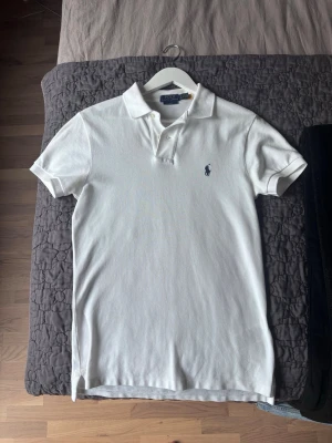 Vit pikétröja från Polo Ralph Lauren - Klassisk vit pikétröja från Polo Ralph Lauren i slim fit-modell. Tröjan har korta ärmar, krage och knappar framtill samt den ikoniska broderade loggan på bröstet. Tillverkad i bomull för en bekväm känsla och stilren look.